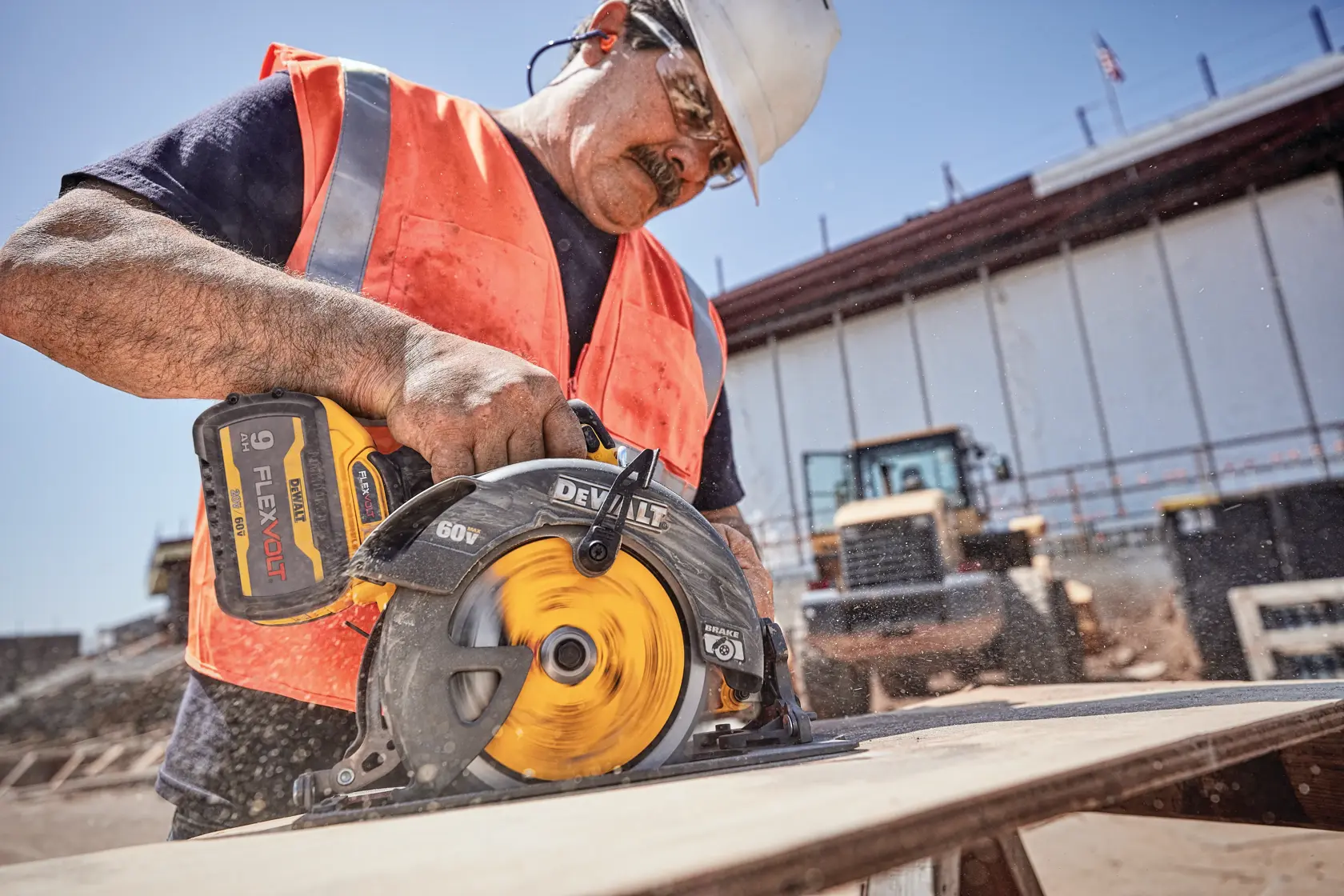 Un trabajador de la construcción con chaleco y casco de seguridad utiliza una sierra circular inalámbrica DEWALT DCS578X2 60V FLEXVOLT para cortar una tabla de madera en una obra al aire libre. La sierra está en funcionamiento, generando polvo de madera visible.