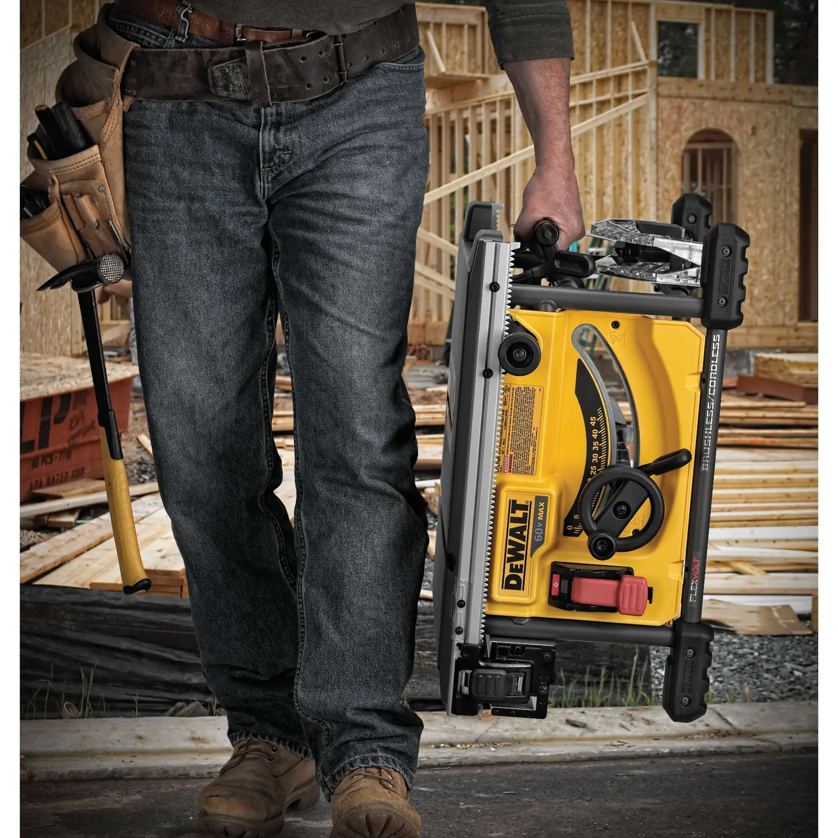 Una persona con jeans de trabajo y botas lleva una sierra de mesa portátil DEWALT en un sitio de construcción, con un cinturón de herramientas y un martillo visibles. Al fondo se observa el armazón de madera de un edificio.