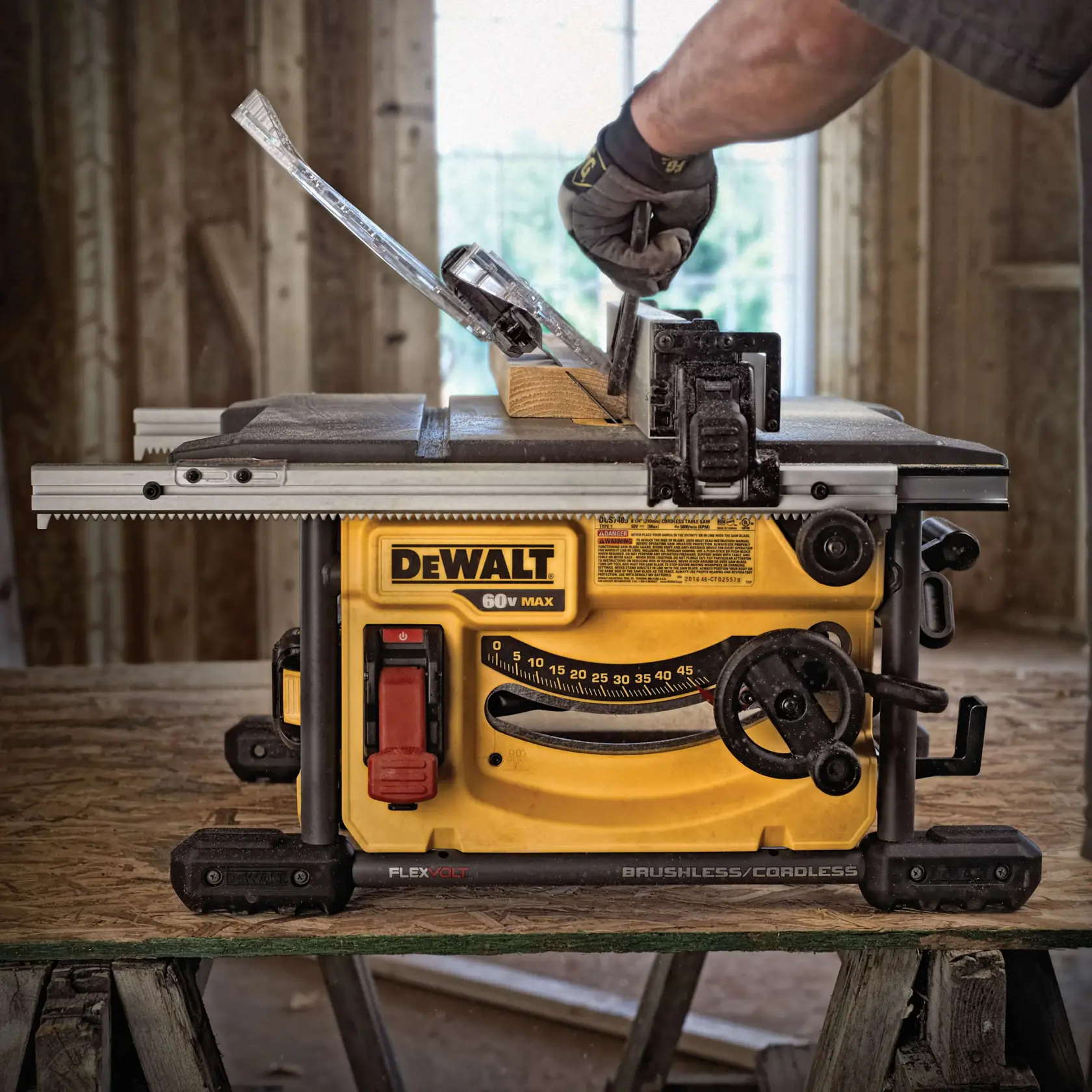 Una sierra de mesa inalámbrica DEWALT 60V Max de color amarillo y negro está colocada sobre una superficie de madera, con una mano enguantada ajustando la herramienta y una pieza de madera lista para cortar. La sierra muestra marcas de medición y perillas de ajuste visibles.
