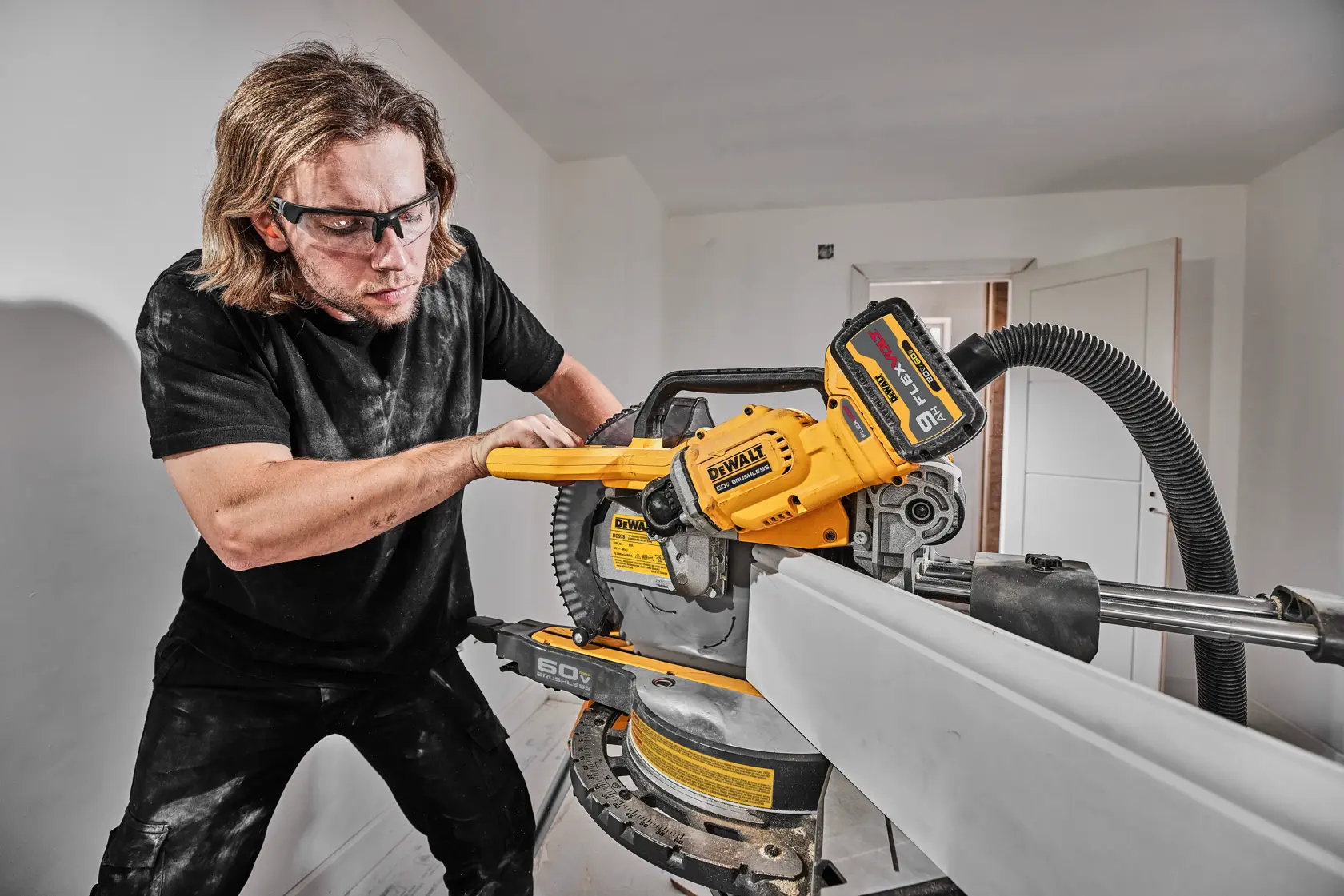 Una persona utiliza una sierra ingletadora inalámbrica DEWALT (modelo DCS781B) en interiores, cortando una pieza larga de material. La herramienta está conectada a una manguera de extracción de polvo. El rostro de la persona está difuminado por privacidad.