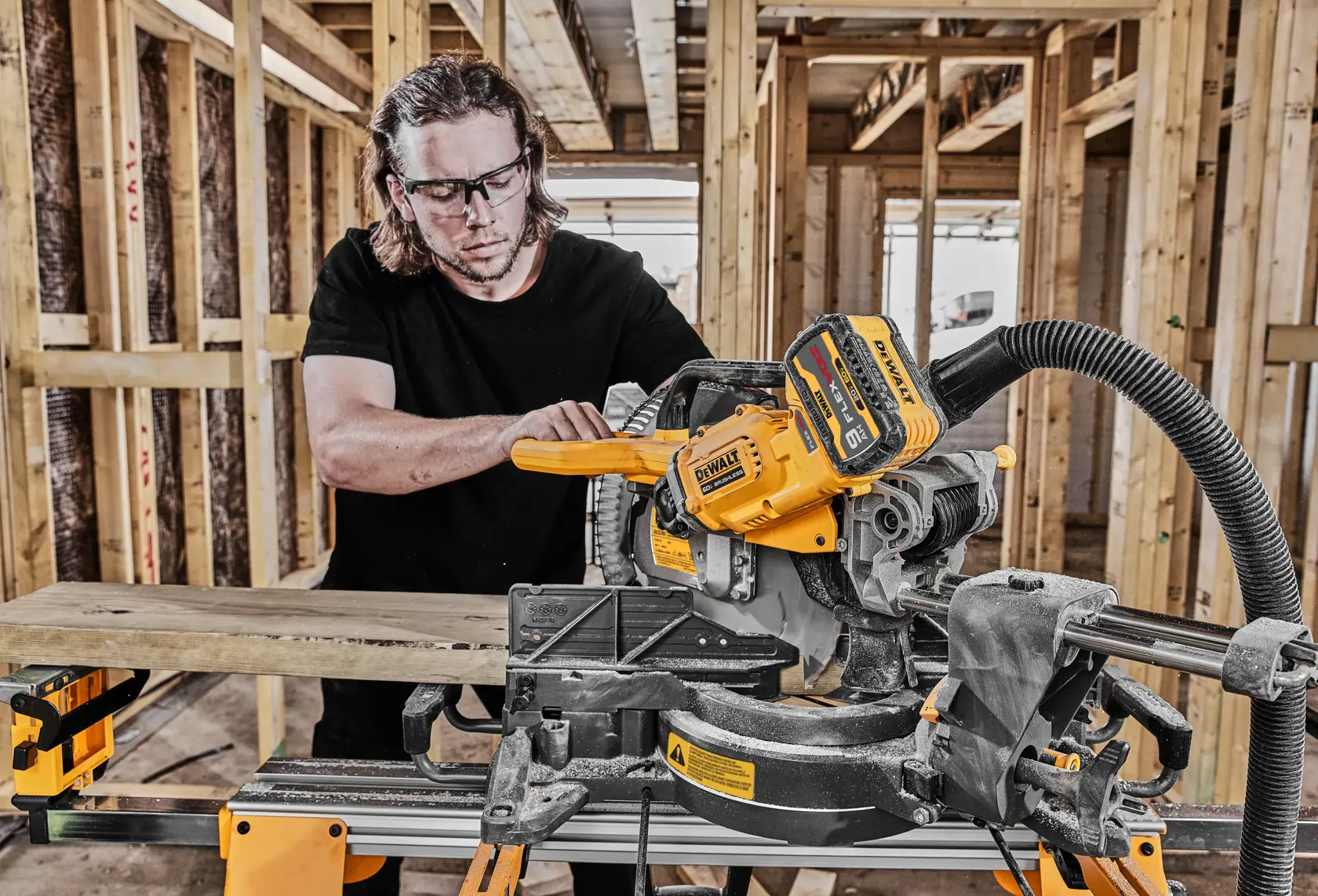 Una persona utiliza una sierra ingletadora telescópica DEWALT para cortar una pieza de madera en un sitio de construcción con estructuras de madera expuestas. La sierra está equipada con batería y una manguera de recolección de polvo.