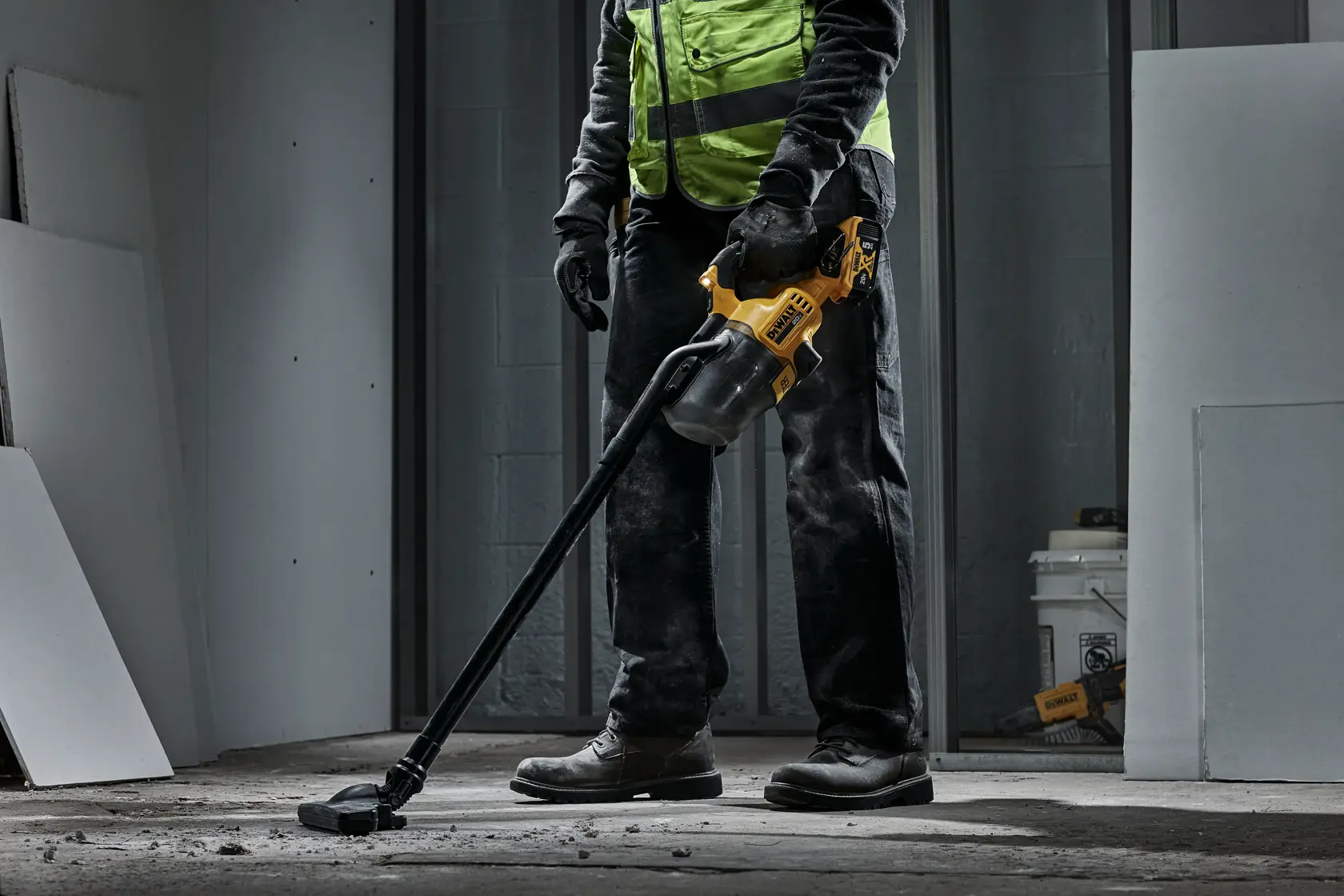 Una persona con chaleco de seguridad de alta visibilidad y guantes usa una aspiradora inalámbrica amarilla y negra DEWALT para limpiar polvo y residuos de un suelo de concreto en una habitación sin terminar.