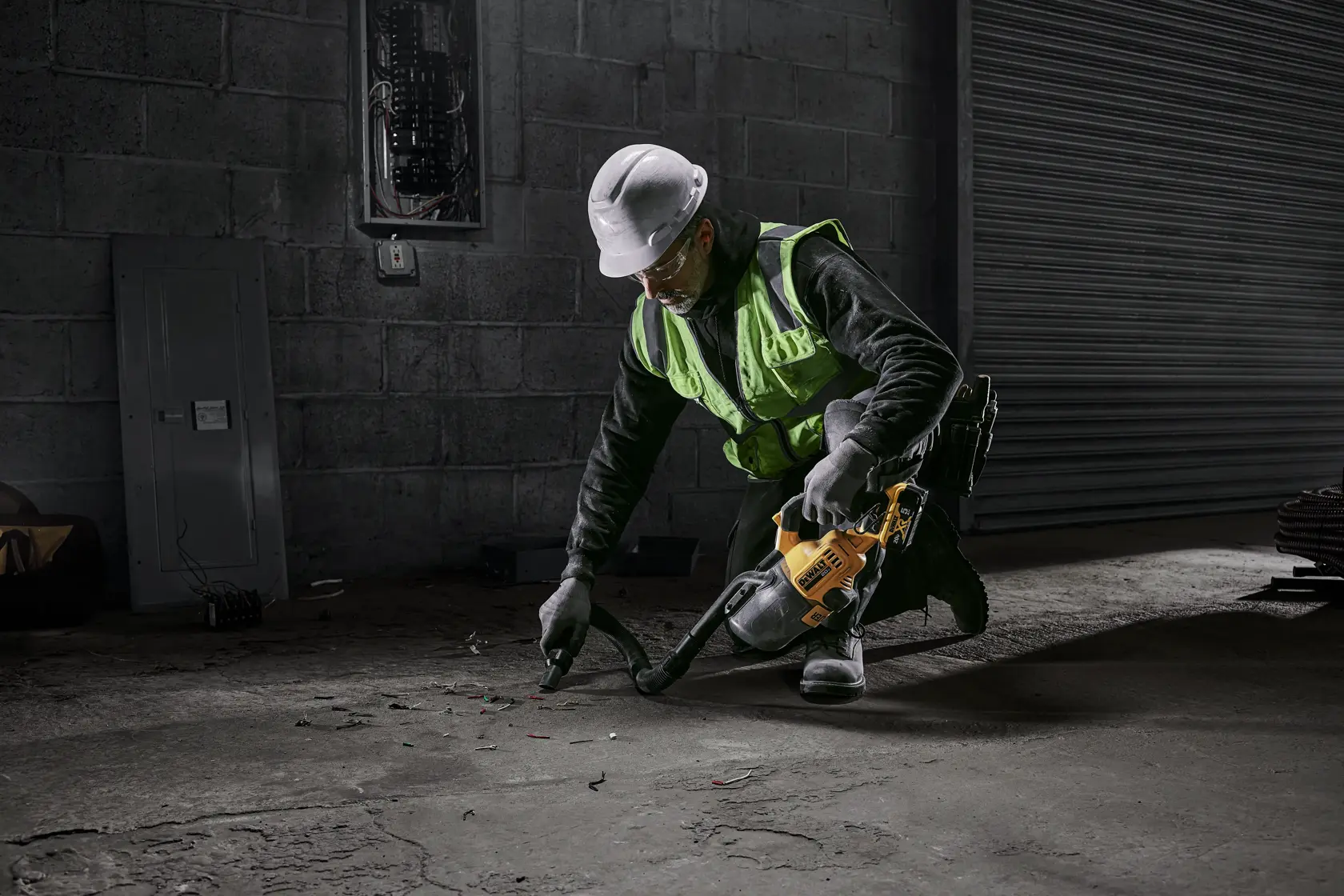 Un trabajador con chaleco de seguridad y casco está arrodillado y usando una aspiradora inalámbrica amarilla DEWALT para limpiar residuos del piso de concreto en un entorno industrial.