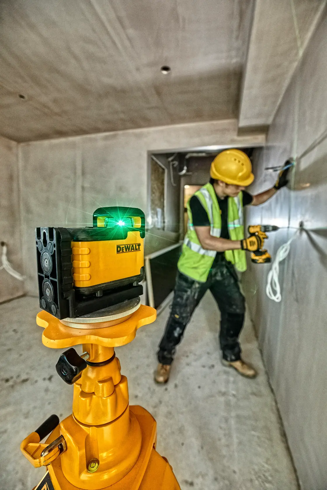Un nivel láser verde DEWALT está colocado sobre un trípode en una habitación en renovación, proyectando una línea verde sobre la pared. Se ve a una persona con el rostro desenfocado en el fondo, cerca de un gabinete y área de trabajo.