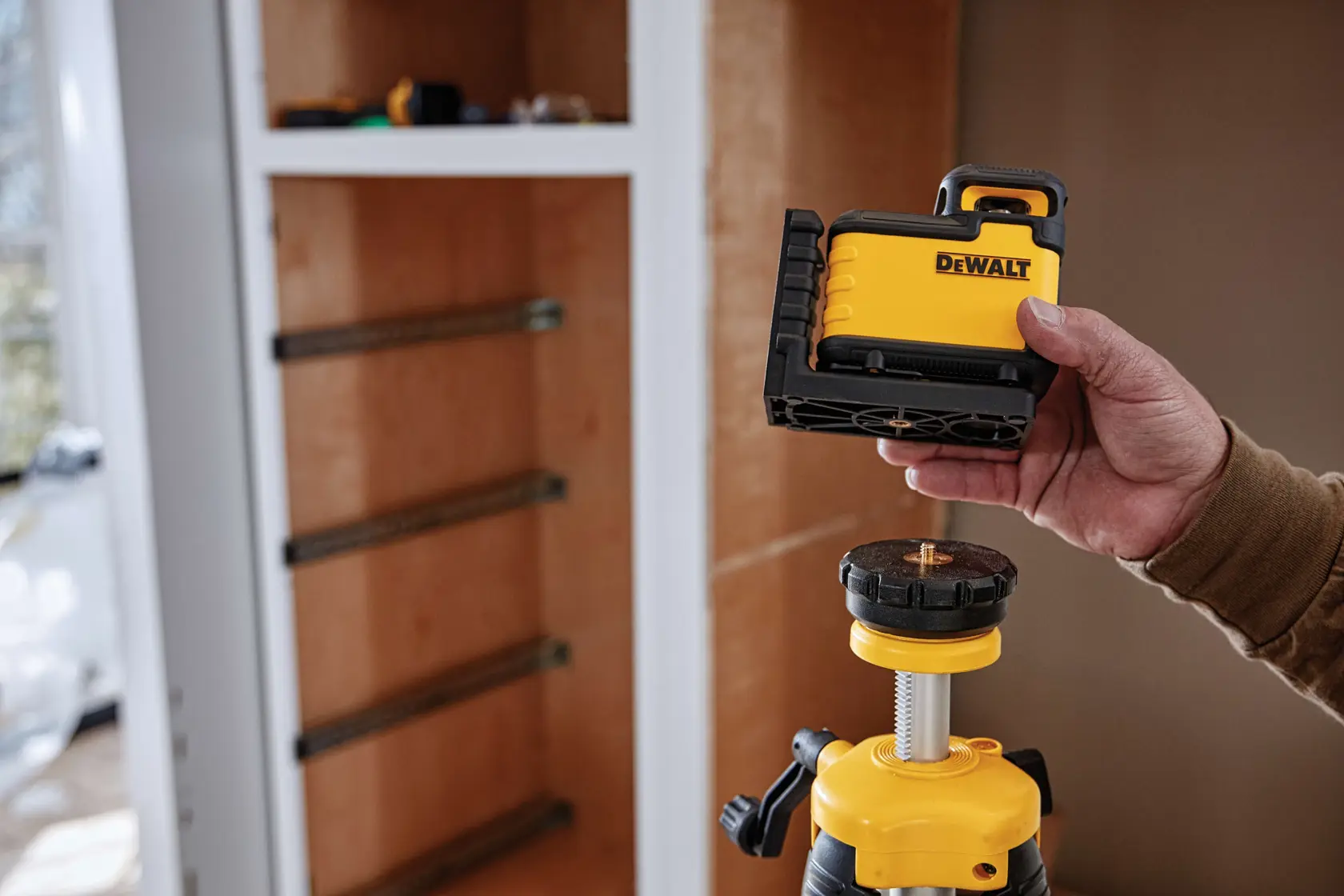 Una persona sostiene una herramienta de nivel láser DEWALT sobre un trípode frente a un gabinete de madera en construcción.