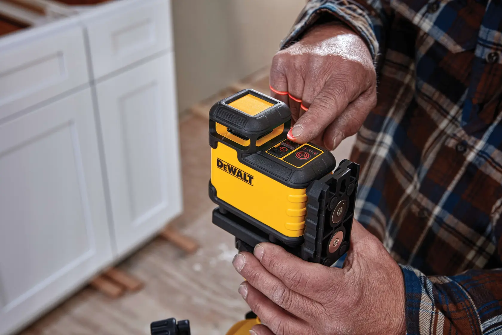 Una persona opera un nivel láser amarillo y negro DEWALT, presionando un botón en la parte superior. El dispositivo está montado sobre un trípode y se utiliza para nivelación y alineación precisa en construcción o renovación. Se observan gabinetes blancos y un piso de madera en el fondo.