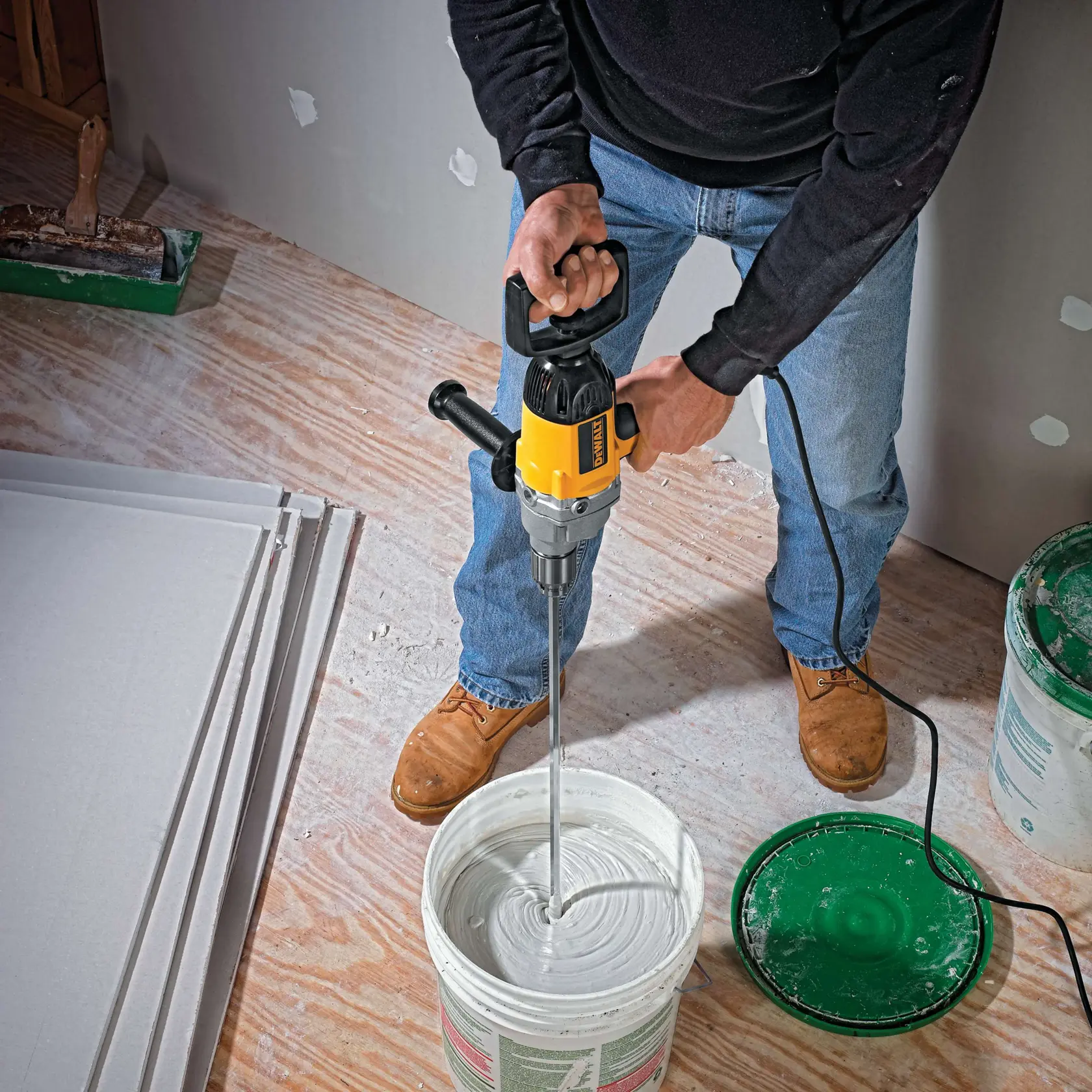 Una persona usa un mezclador eléctrico DEWALT DW130V para mezclar pintura o yeso en un gran balde sobre un suelo de madera. Se pueden ver hojas de yeso y materiales de construcción cerca.
