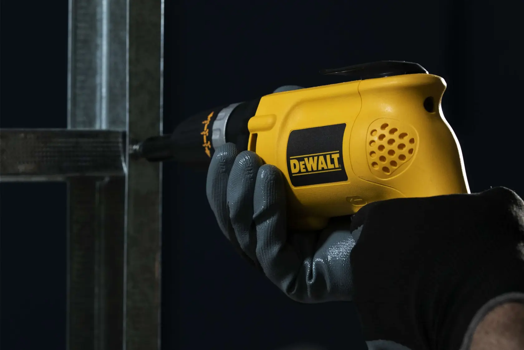 Primer plano de una mano enguantada sosteniendo una herramienta eléctrica amarilla DEWALT, utilizada para fijar un tornillo en un marco de metal.