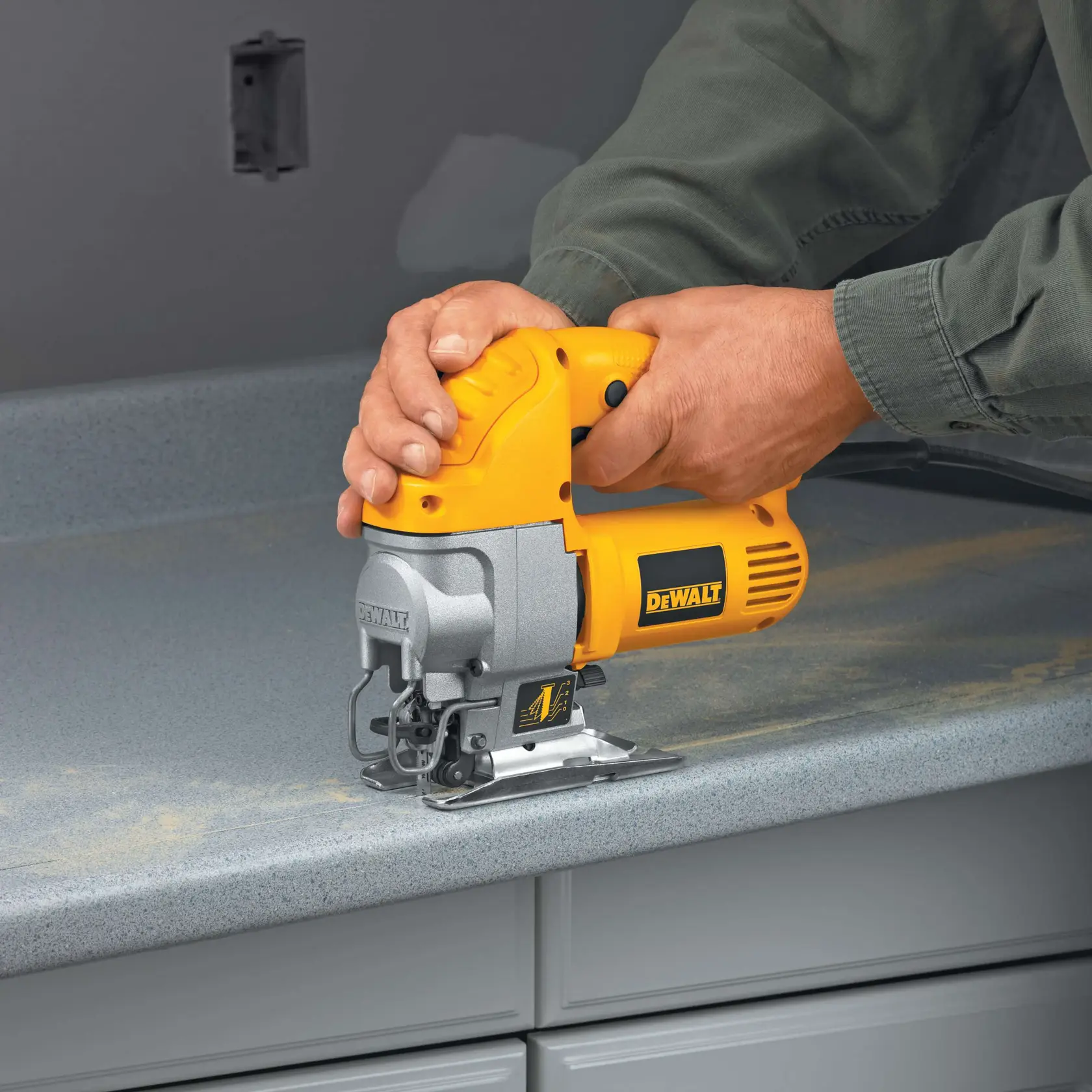 Una persona usa una sierra de calar DEWALT amarilla y plateada para cortar una encimera gris. La herramienta está sujetada con firmeza y se puede ver polvo de madera en la superficie.