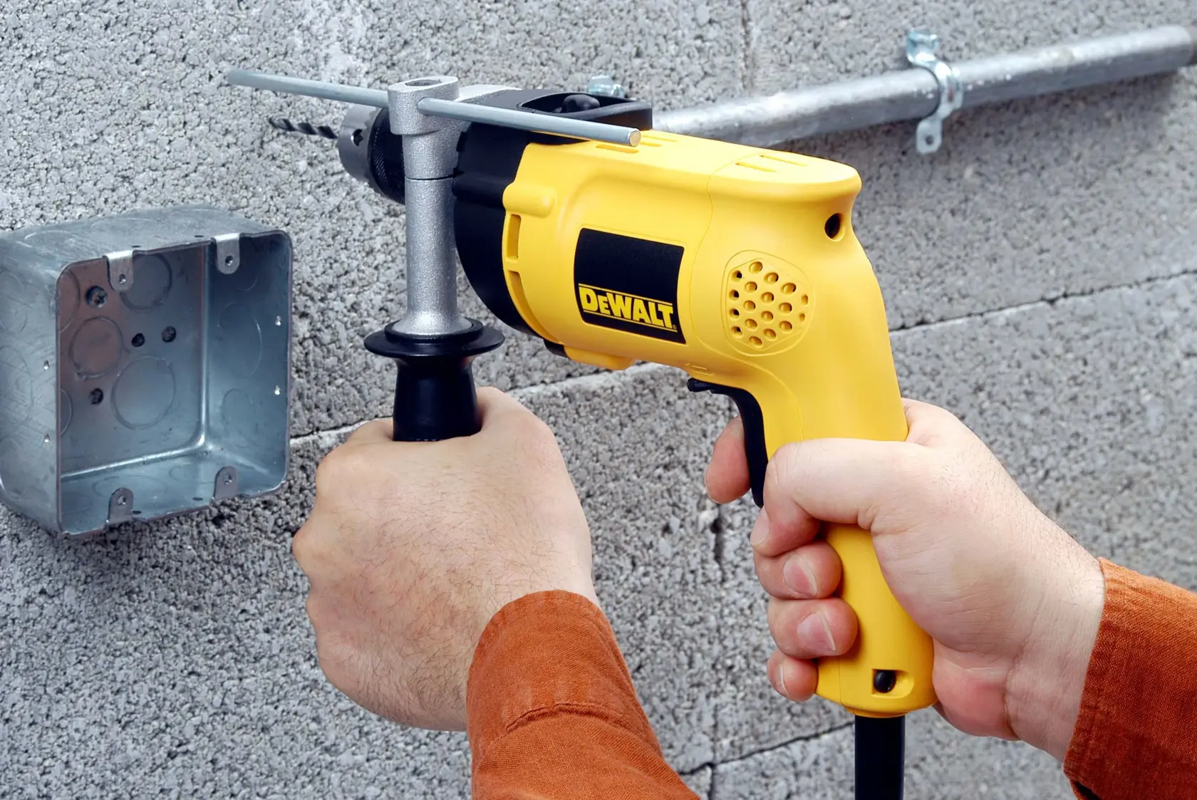 Una persona está usando un taladro eléctrico amarillo DEWALT para hacer un agujero en una pared de hormigón cerca de una caja eléctrica de metal.