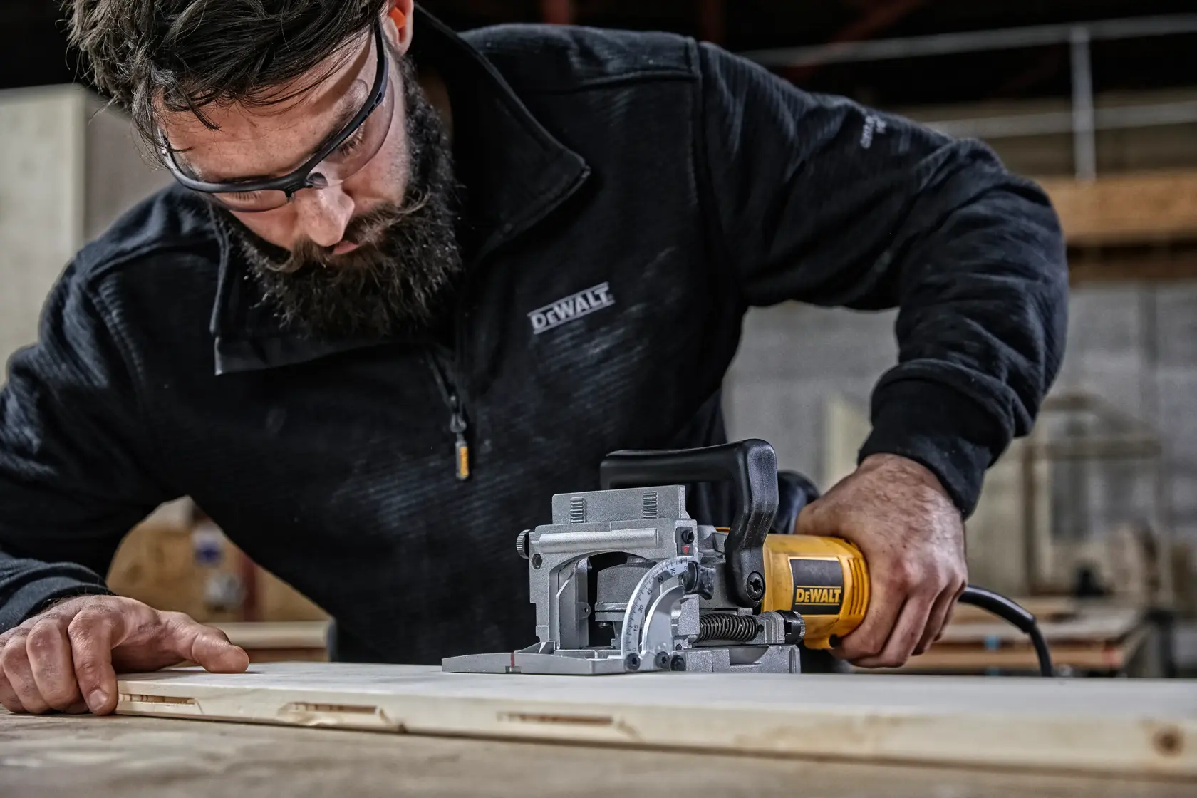 Una persona utiliza una fresadora para unión de galleta DEWALT DW682K sobre una superficie de madera en un taller. El rostro del usuario está desenfocado y lleva una chaqueta negra DEWALT.