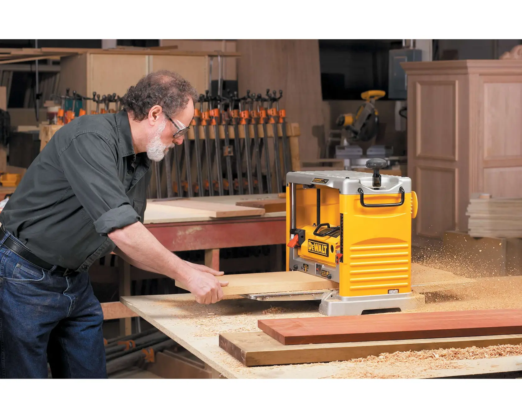 Una persona operando una cepilladora de madera DEWALT DW734 en un taller, alisando un trozo de madera mientras las virutas de madera vuelan alrededor. El espacio de trabajo está equipado con abrazaderas, mesas y tablas de madera.
