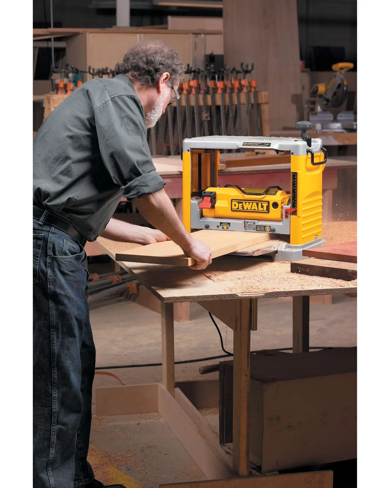 Una persona usando una cepilladora de madera DEWALT DW734 sobre un banco de trabajo en un taller de carpintería. La cepilladora se utiliza para alisar una tabla de madera, con virutas de madera visibles en la mesa.