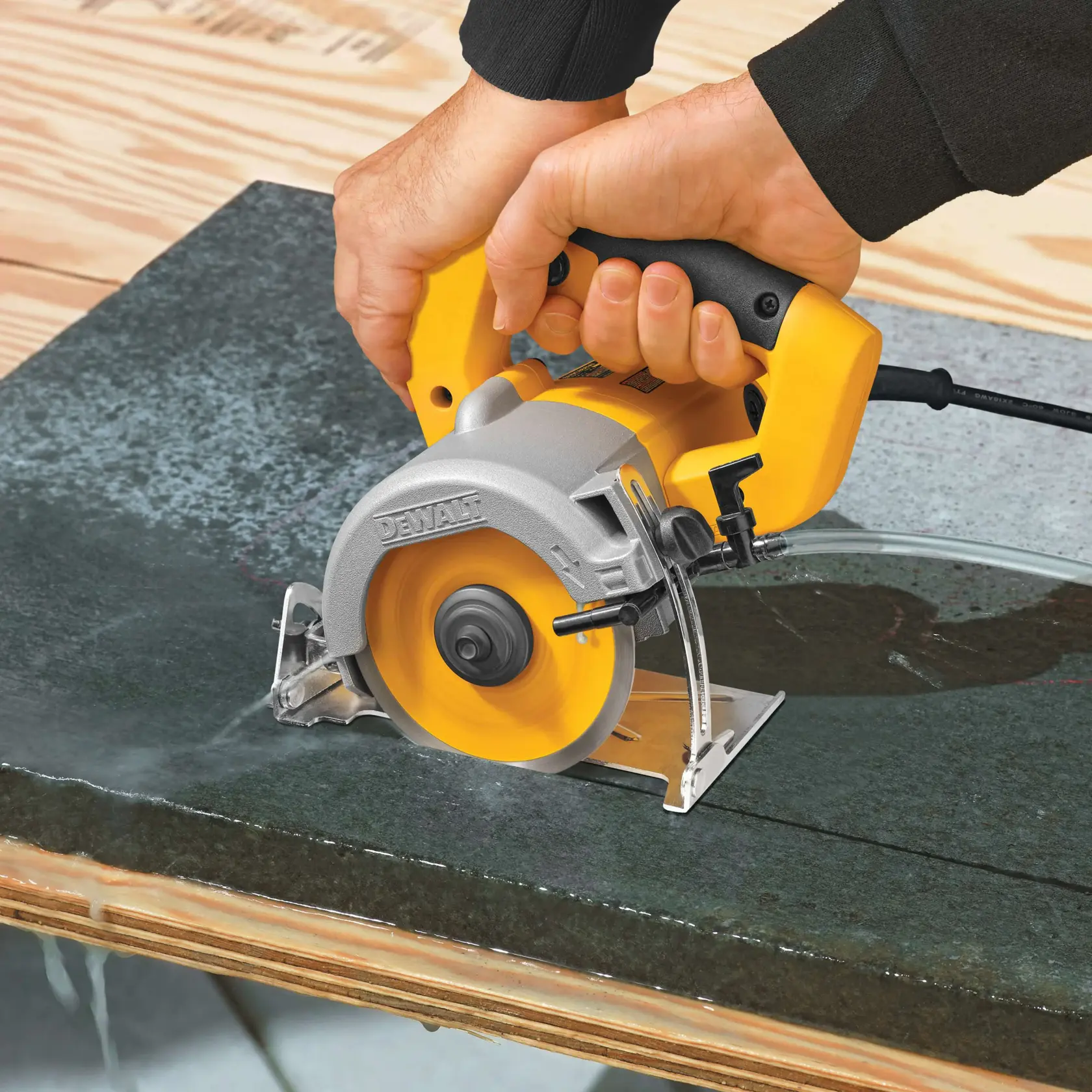 Una persona utiliza una sierra para azulejos húmedos DEWALT para cortar una pieza de piedra o azulejo oscuro sobre una superficie de trabajo de madera. Se utiliza agua para enfriar la hoja.