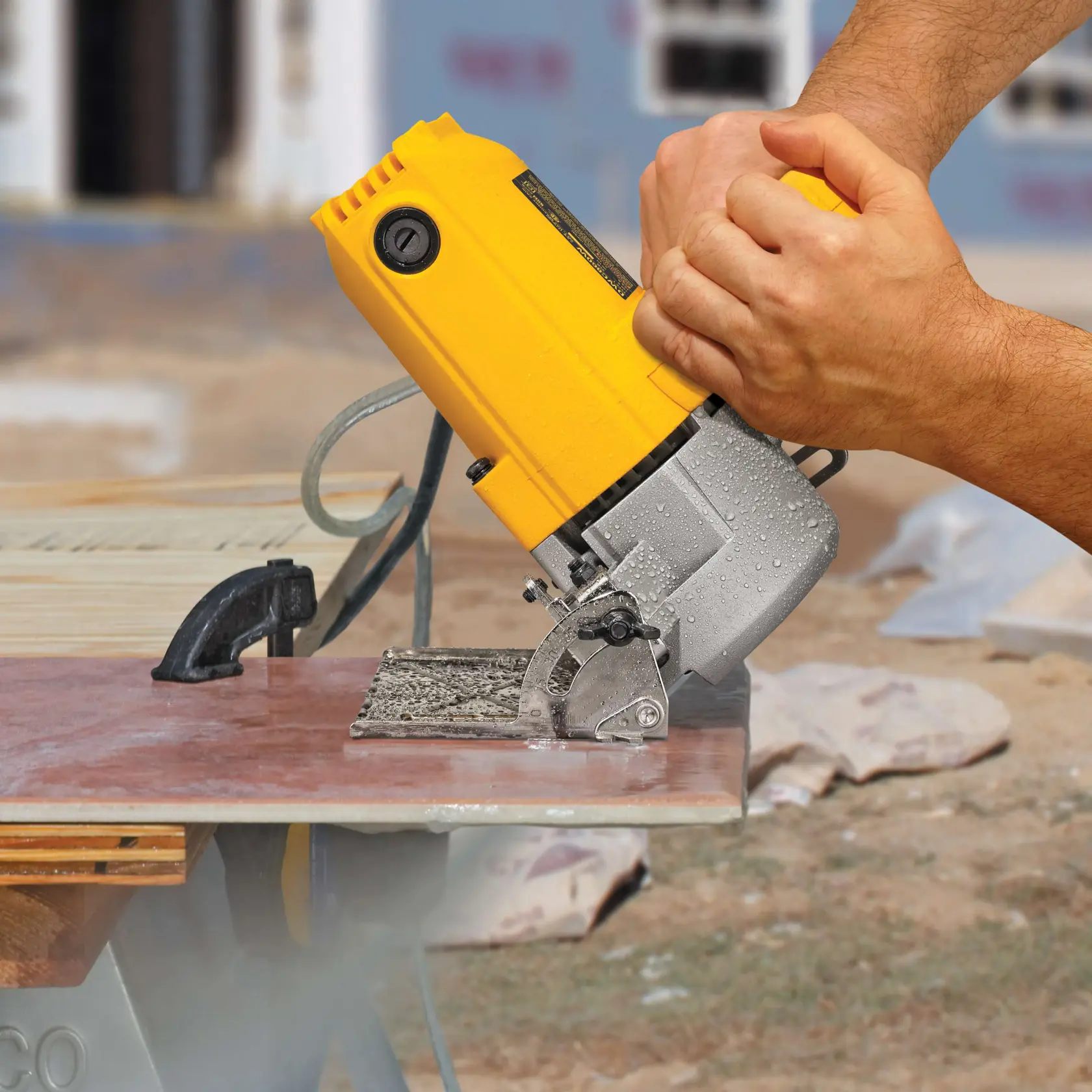 Un primer plano de unas manos operando una cortadora de azulejos amarilla DEWALT, cortando una baldosa de cerámica en un banco de trabajo al aire libre.
