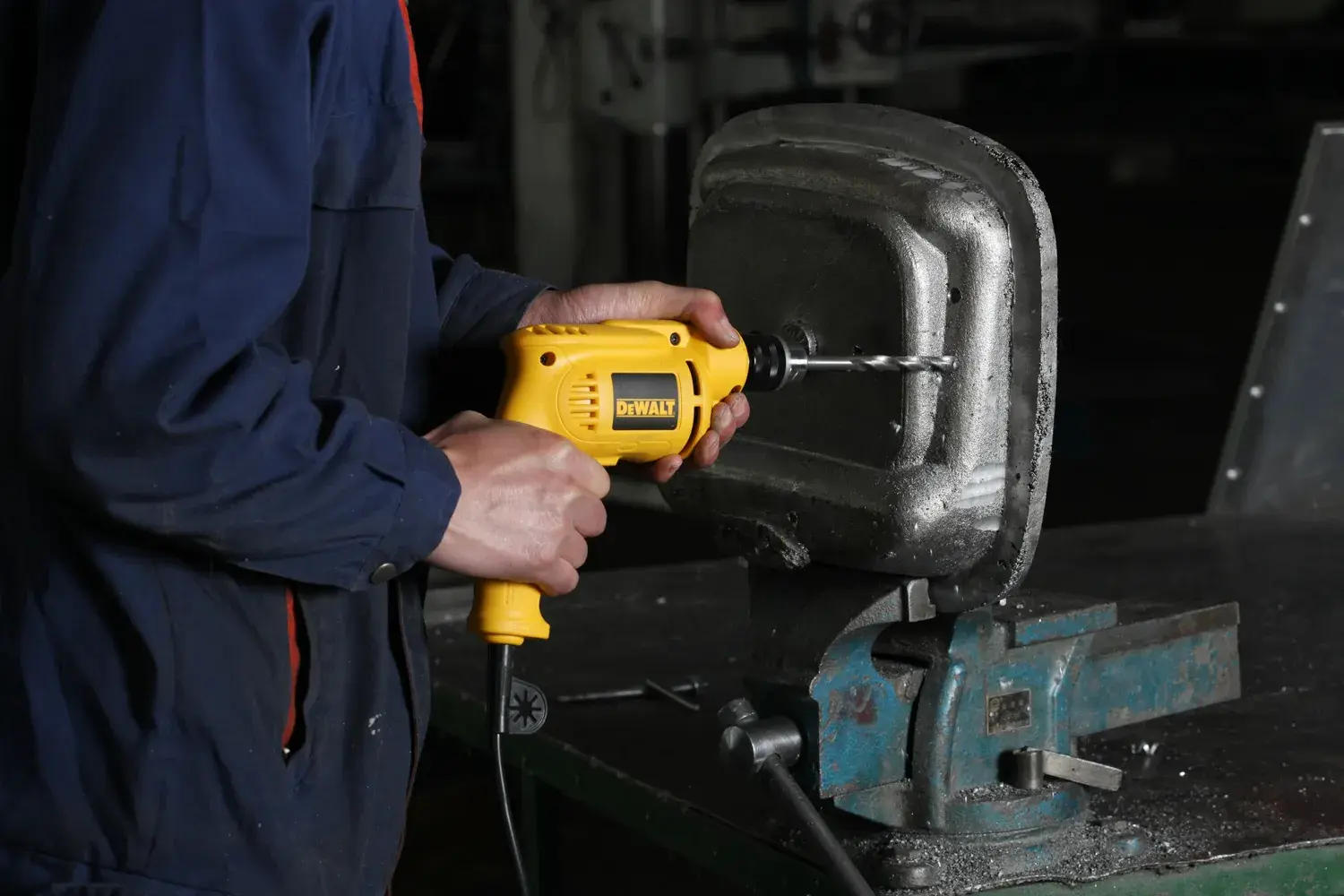 Una persona utiliza un taladro eléctrico amarillo DEWALT para trabajar en un objeto metálico sujetado en una prensa sobre un banco de trabajo.