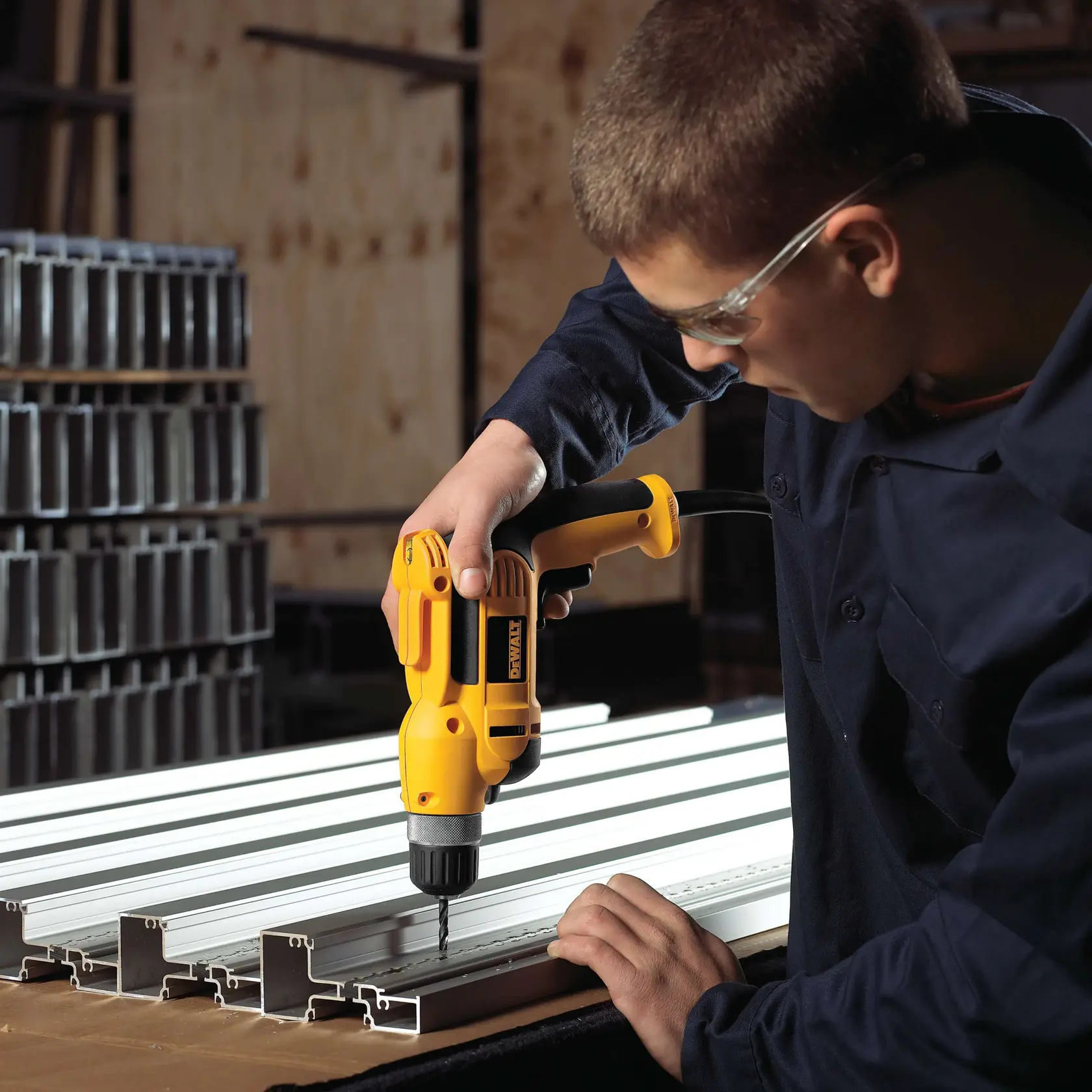 Una persona utiliza un taladro eléctrico amarillo DEWALT para perforar perfiles metálicos colocados sobre una mesa de trabajo. La persona lleva una camisa de trabajo oscura y gafas de seguridad. El fondo incluye pilas de secciones metálicas y un entorno de taller.
