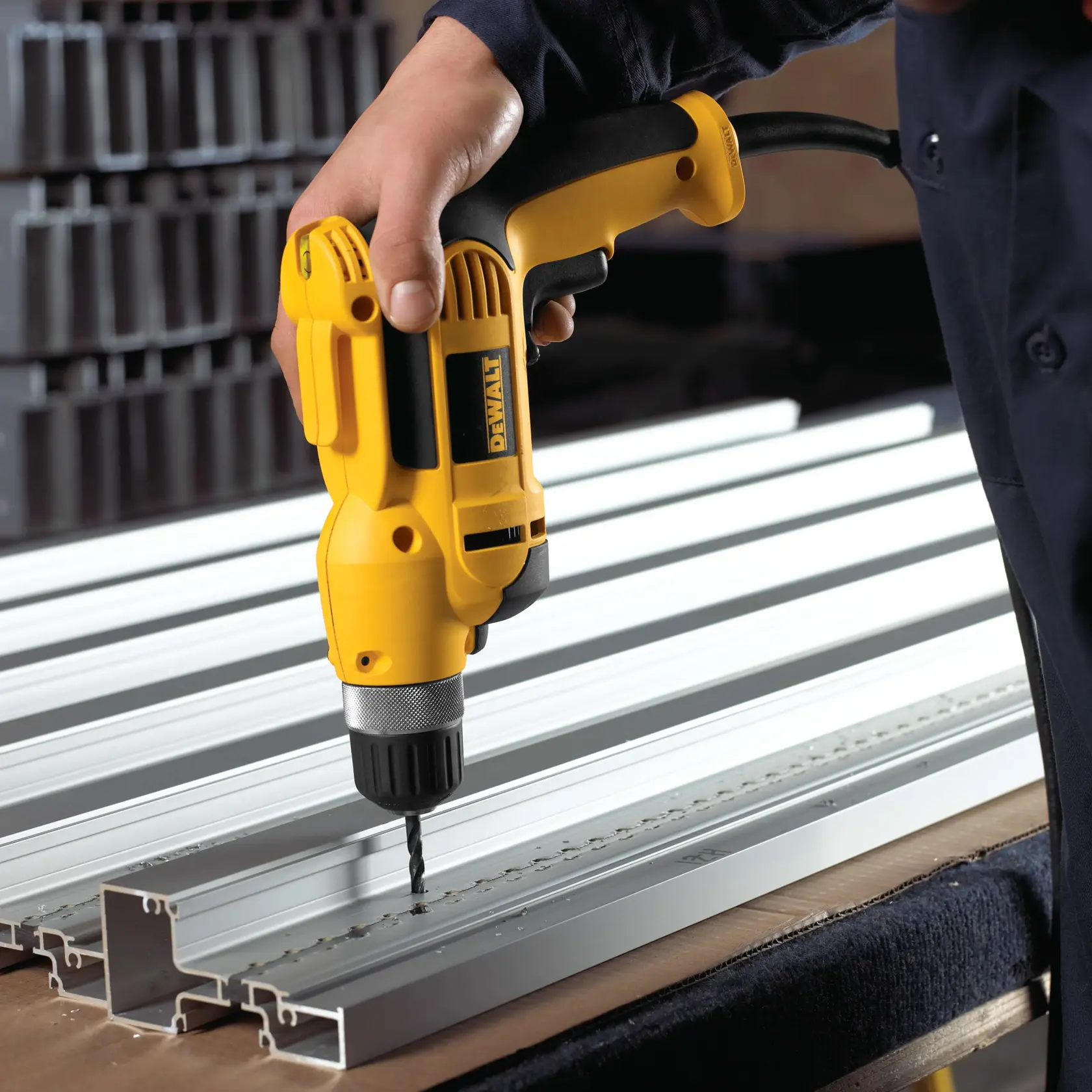 Una persona está utilizando un taladro eléctrico amarillo DEWALT para hacer agujeros en canales de metal de aluminio colocados en una mesa de trabajo.