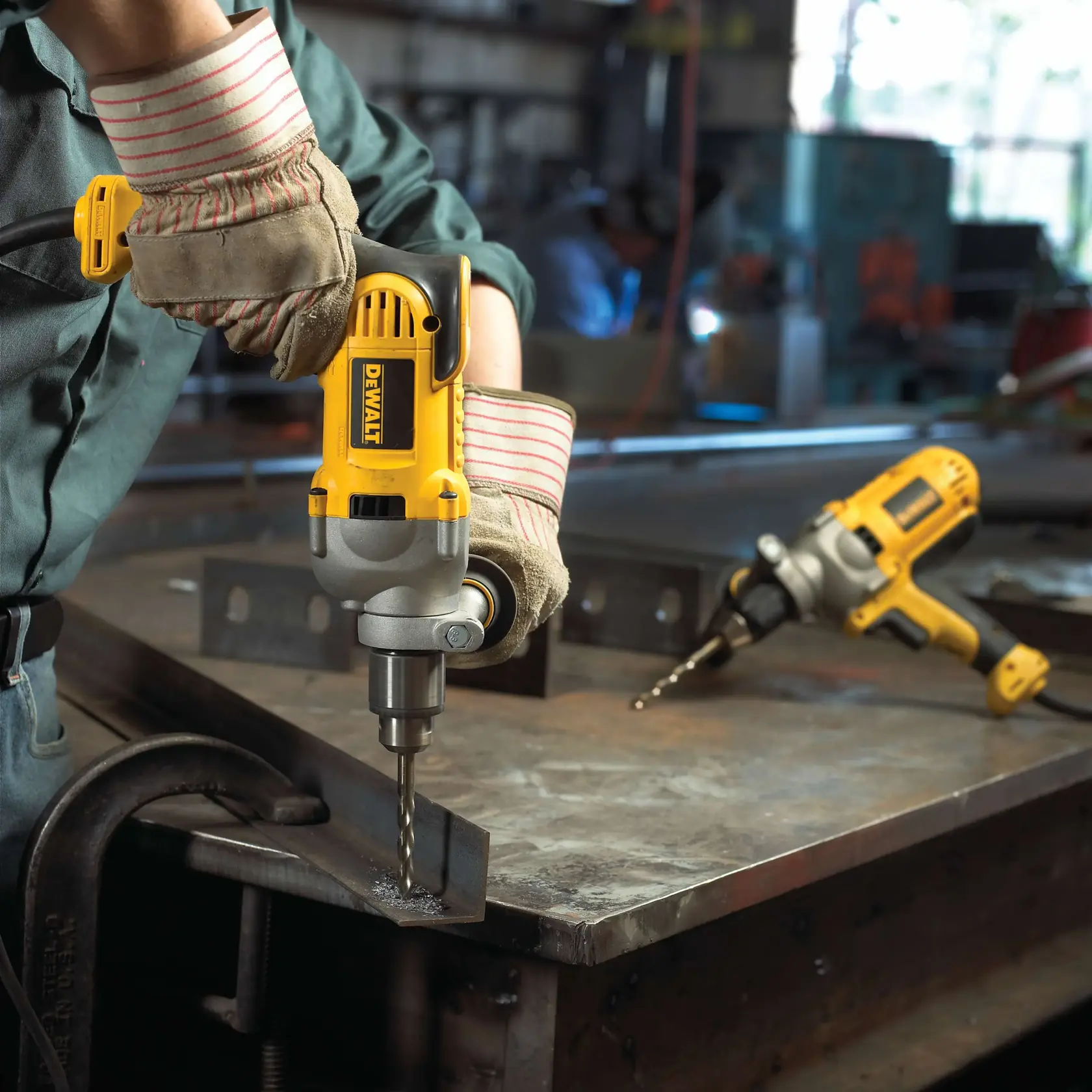 Una persona con guantes de trabajo está usando un taladro eléctrico amarillo DEWALT para perforar metal sobre un banco de trabajo. Otro taladro DEWALT está apoyado en el banco en el fondo. La escena parece estar en un taller.