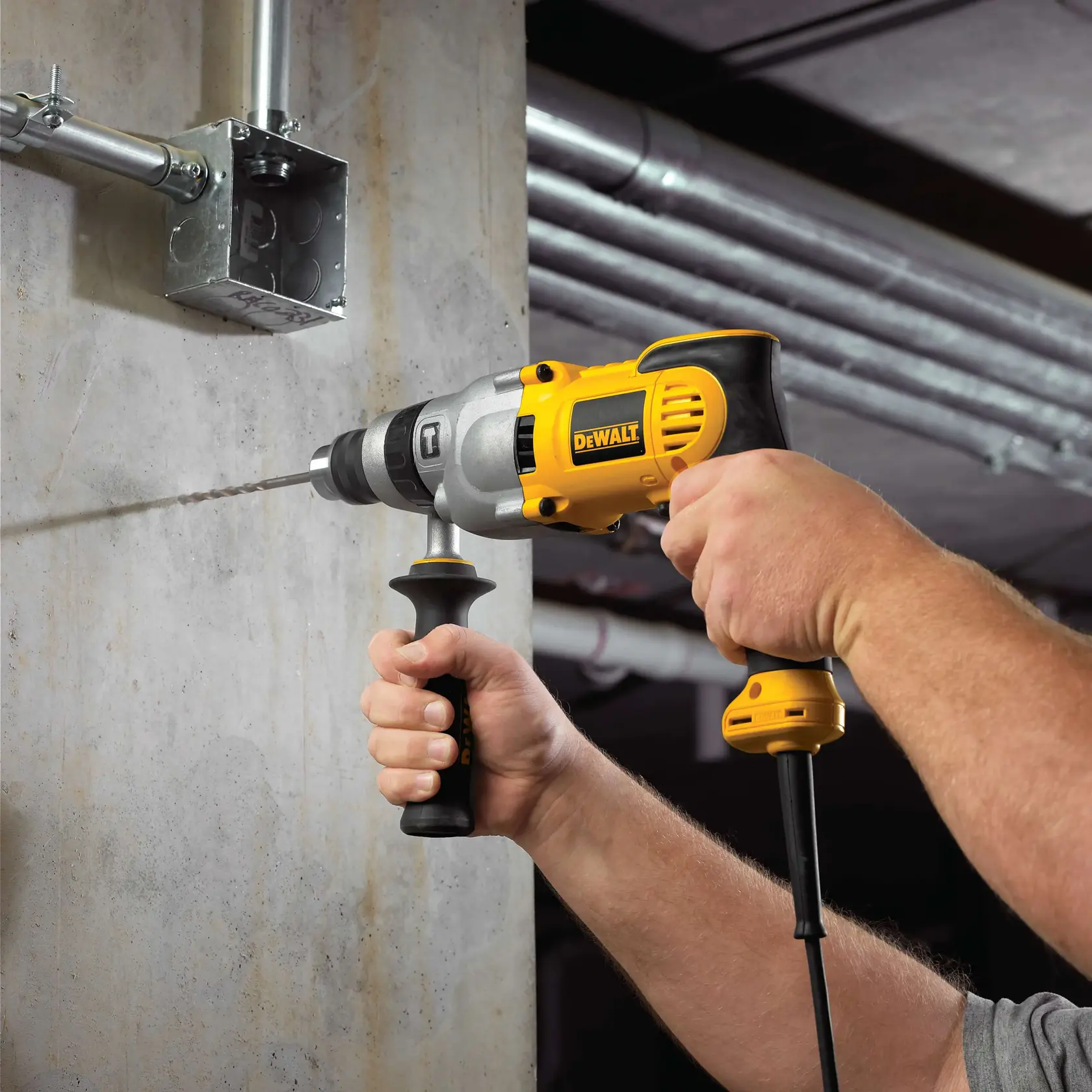 Una persona utiliza un taladro eléctrico con cable DEWALT con una empuñadura lateral para perforar una pared de hormigón. El taladro es amarillo y negro y se utiliza en un entorno industrial.