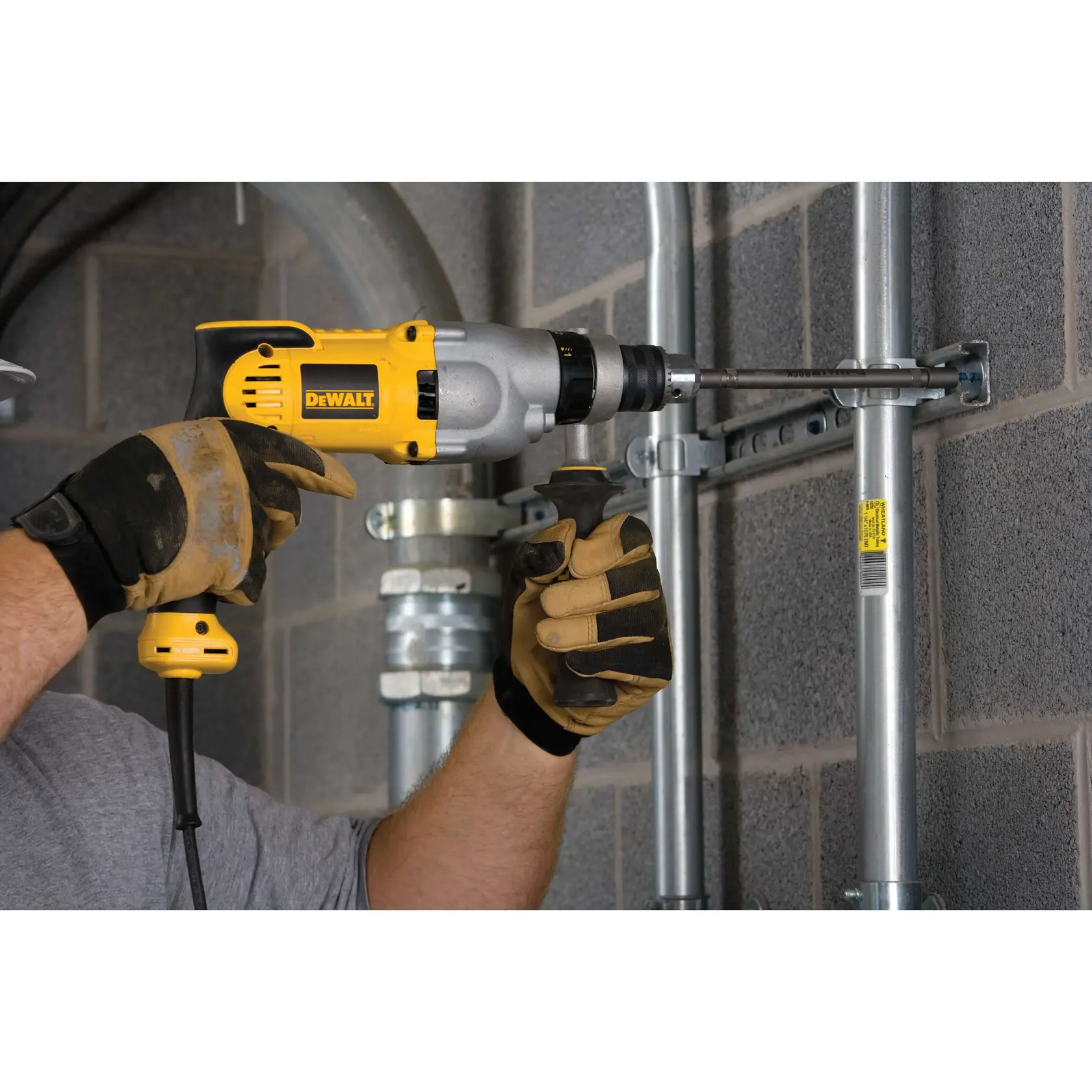 Una persona con guantes de trabajo está usando un taladro eléctrico amarillo y negro DEWALT para instalar o asegurar un conducto metálico contra una pared de bloques de hormigón gris en un entorno industrial.