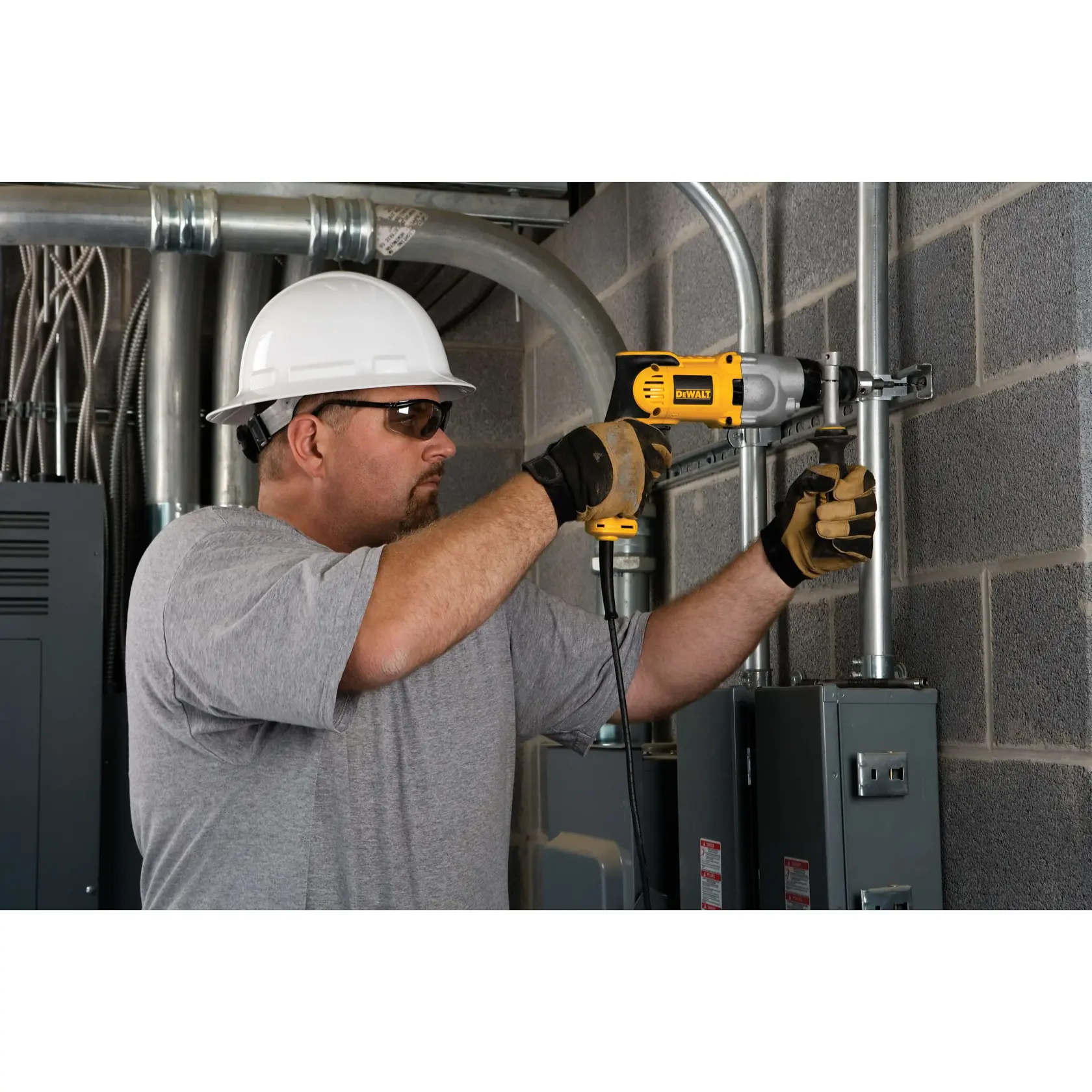Una persona con casco blanco y guantes está usando una taladradora DEWALT DWD520 para trabajar en tubos de metal sujetos a una pared de ladrillo en un entorno industrial.