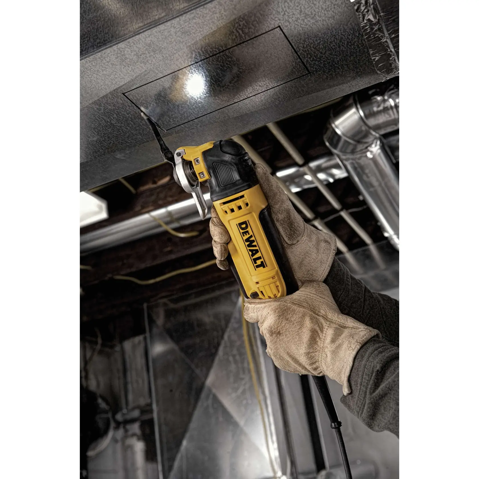 Una persona con guantes de trabajo utiliza una herramienta multifunción oscilante DEWALT DWE315 para cortar un contorno cuadrado en un conducto metálico. La herramienta está encendida y tiene una luz incorporada para mayor precisión. La imagen se toma en un entorno industrial con tubos y conductos visibles.