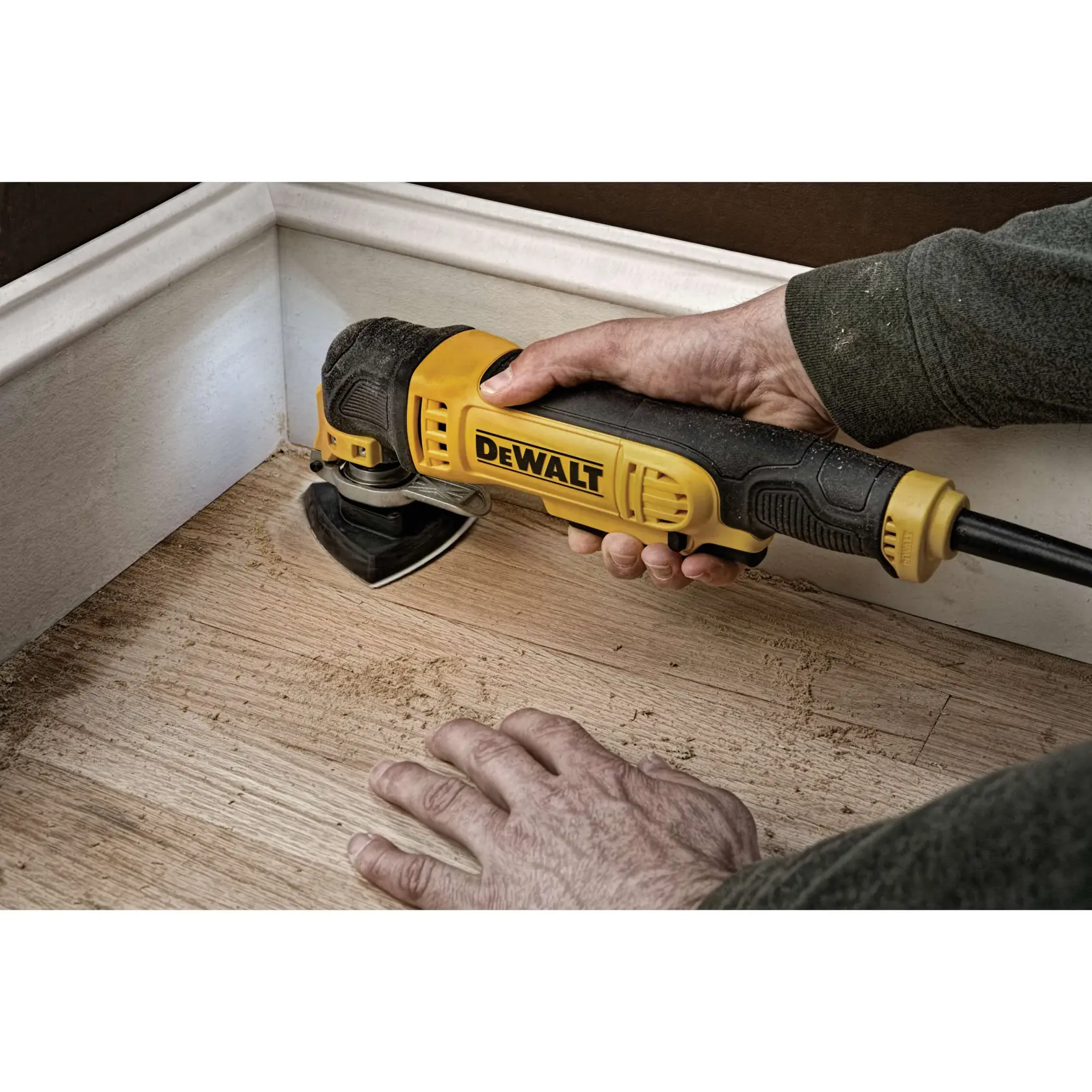 Una persona está usando una herramienta multitarea oscilante DEWALT para lijar o cortar el suelo de madera cerca de un zócalo.