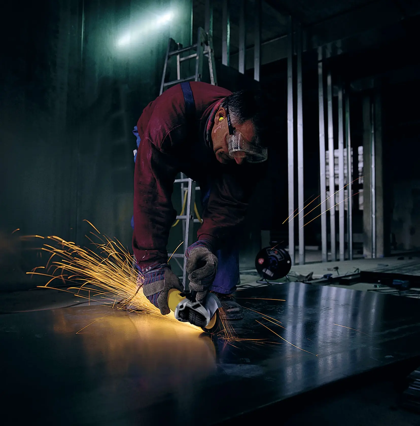 Una persona con guantes protectores y tapones para los oídos utiliza una amoladora angular DEWALT para cortar una hoja de metal, generando chispas en un entorno industrial. El rostro de la persona está difuminado.