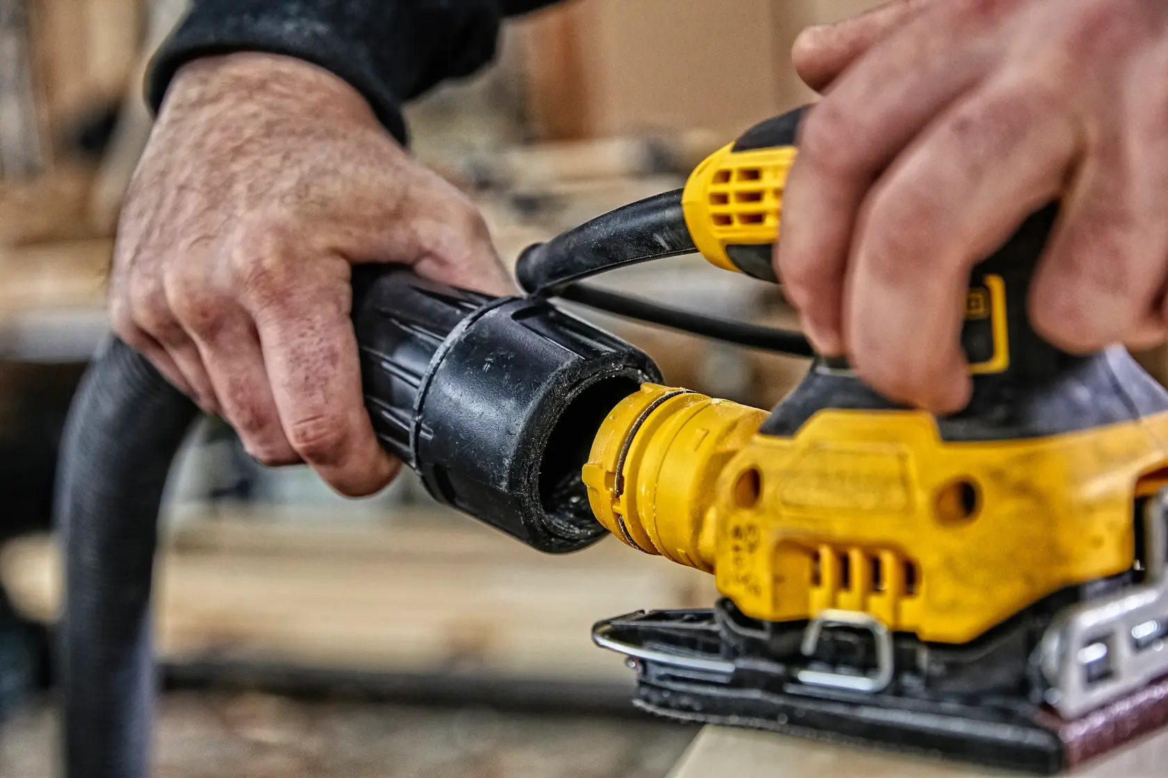 Manos conectan una manguera a una herramienta DEWALT amarilla, usada para trabajar o lijar madera.