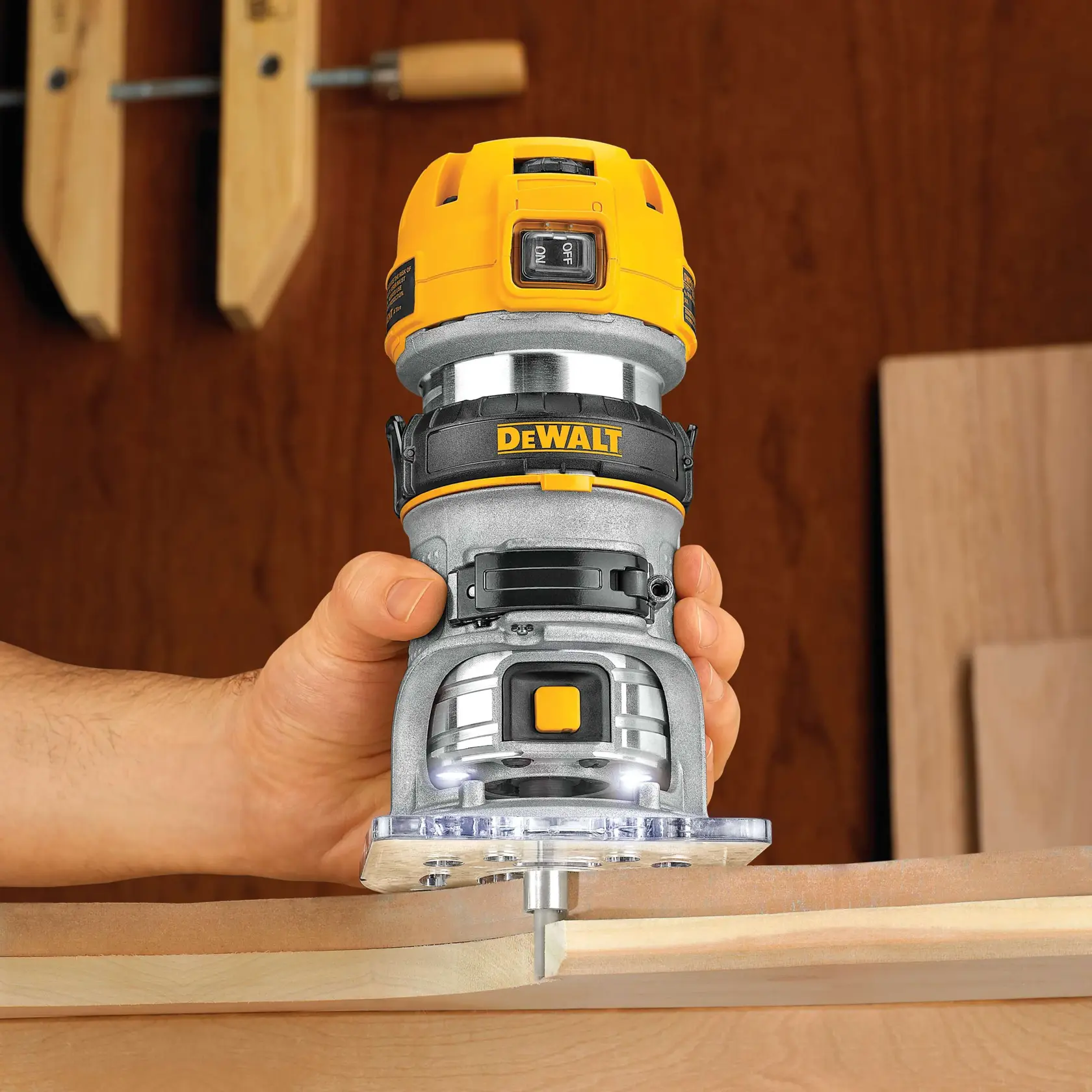 Una mano sostiene una fresadora compacta DEWALT DWP611PK, trabajando en el borde de una pieza de madera. La fresadora es amarilla y plateada, con un botón de encendido visible y luces LED en la base. Hay abrazaderas y tablas de madera en el fondo.