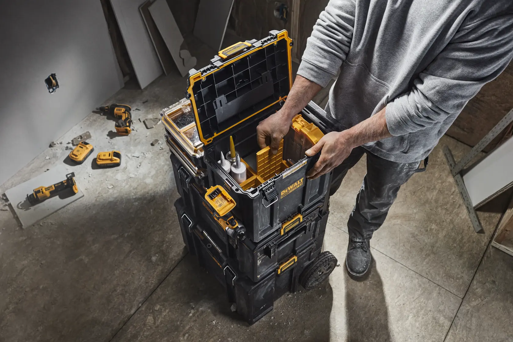 Una persona está organizando herramientas dentro de una caja modular apilada DEWALT en un taller. Otros accesorios y herramientas eléctricas son visibles en el suelo cerca.