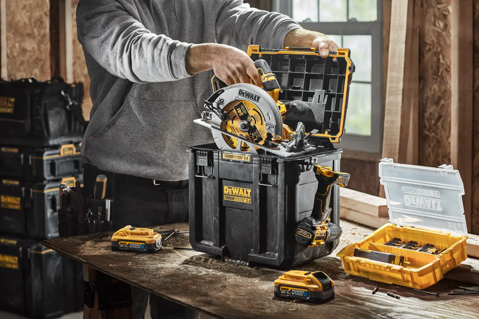 Una caja de almacenamiento DEWALT ToughSystem 2.0 se utiliza para guardar una sierra circular inalámbrica y otras herramientas y accesorios DEWALT sobre una mesa de trabajo dentro de un taller. Hay más cajas y accesorios DEWALT visibles en el fondo.