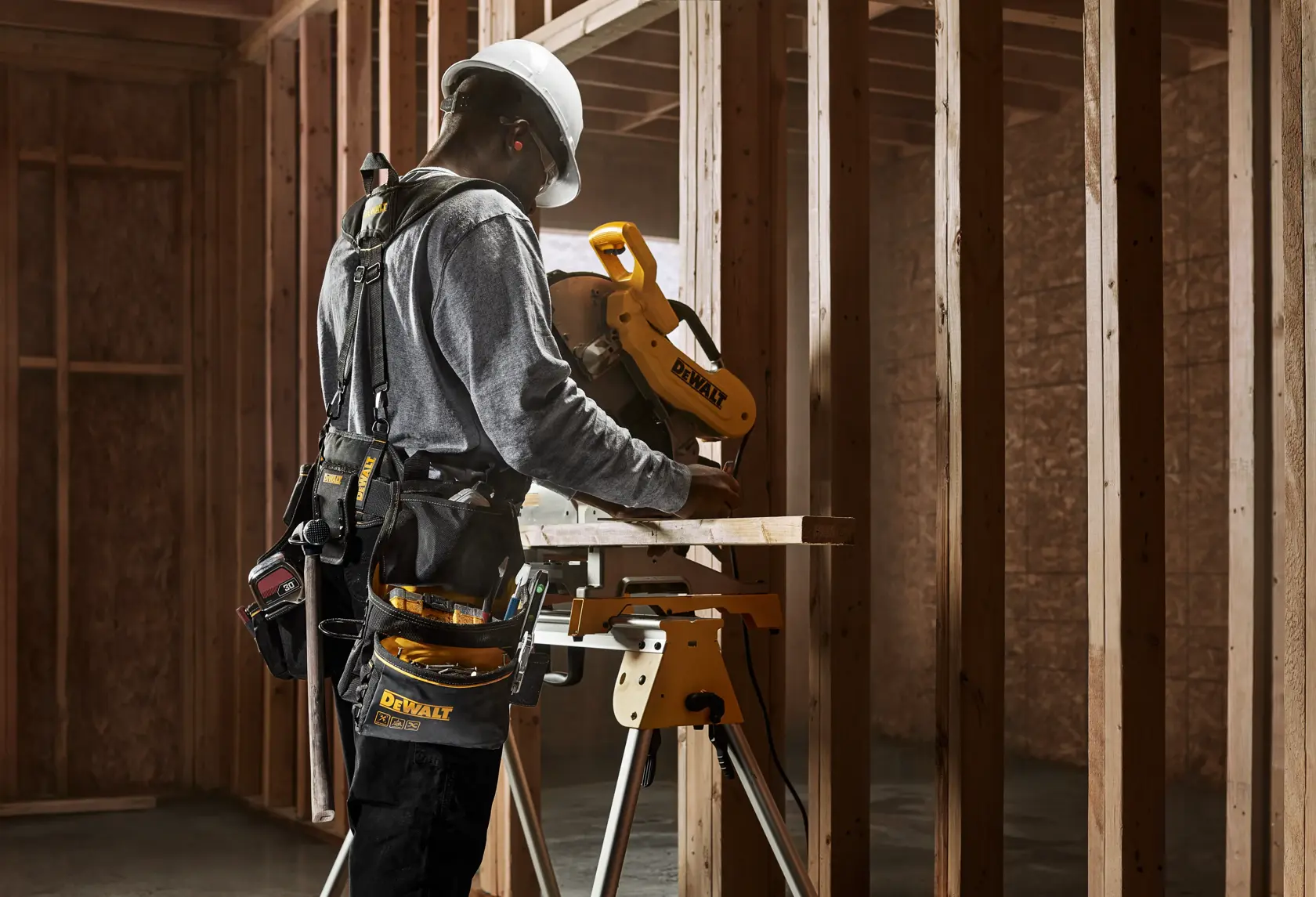 Una persona con casco y cinturón de herramientas utiliza una sierra ingletadora DEWALT en una obra con estructura de madera.
