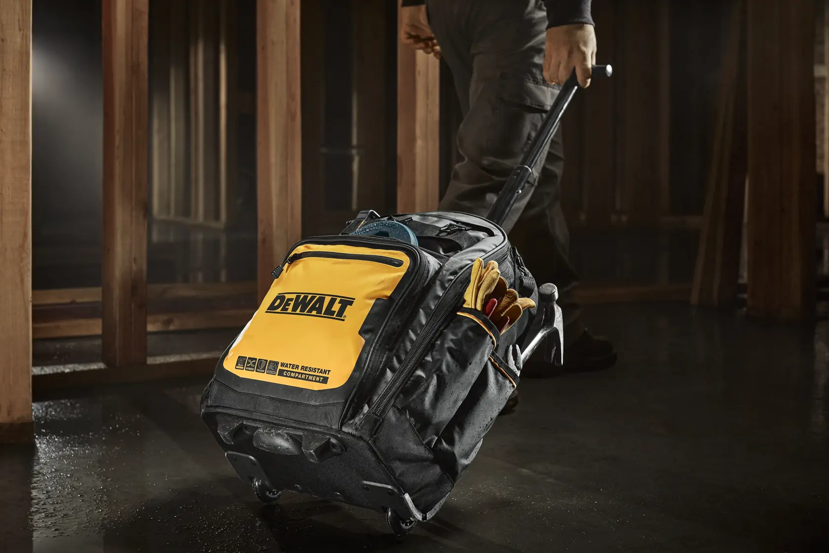 Una bolsa de herramientas rodante DEWALT de diseño amarillo y negro, que es arrastrada por una persona en un entorno de construcción. La bolsa parece ser resistente al agua y contiene varias herramientas y guantes en sus bolsillos laterales.