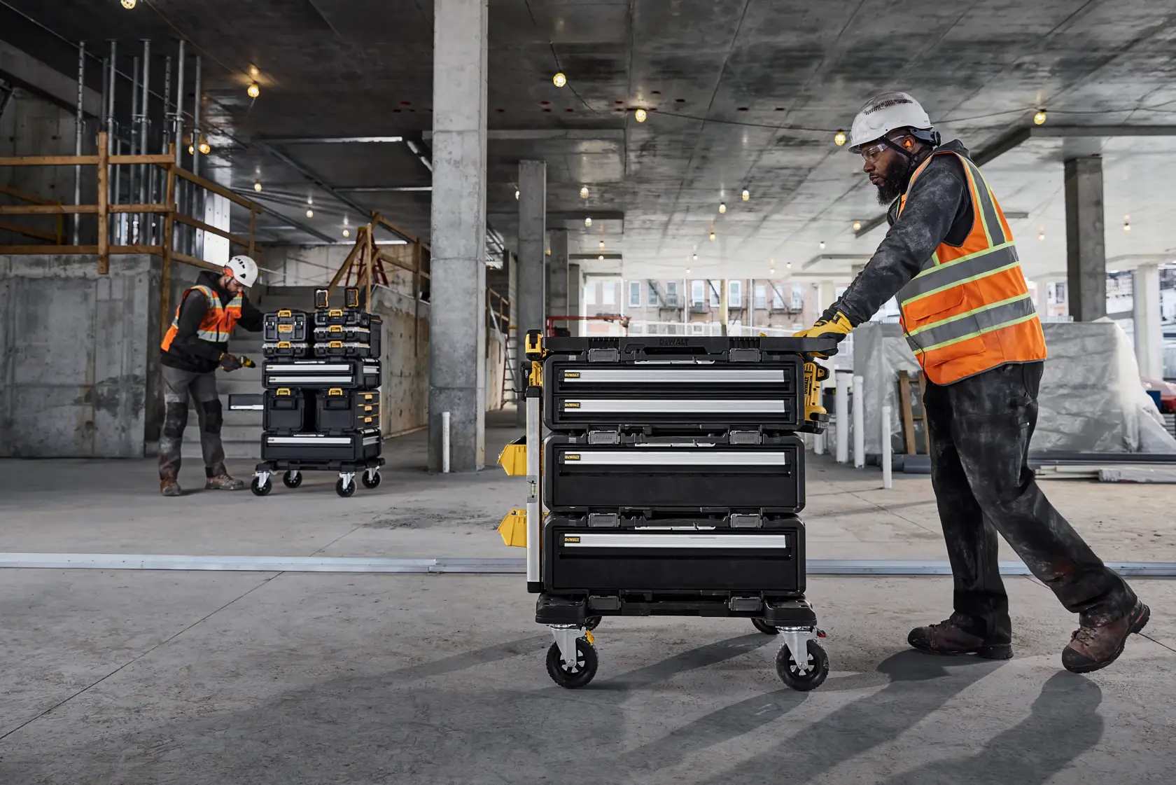 Dos trabajadores con equipo de seguridad y chalecos naranjas utilizan carros de almacenamiento de herramientas portátiles DEWALT con varios cajones sobre ruedas en una gran obra de construcción con columnas de concreto y luces en el techo.